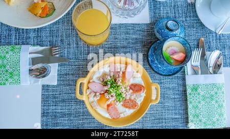 Frühstück mit Spiegeleiern in einer Pfanne mit Speck und Gemüse, Frühstück in einem Luxushotel in Thailand, gebratene Eier in einer kleinen Pfanne mit Schweinehackfleisch und Würstchen Stockfoto