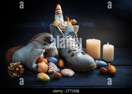 Blaue Stiefel gefüllt mit Süßigkeiten, Weihnachtsdekoration und ein Schokoladen-Weihnachtsmann auf dunklem Holz mit Kerzen, Tradition auf dem deutschen Nikolaus-Tag, was Nikolaus bedeutet Stockfoto