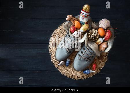 Winterstiefel gefüllt mit einem SchokoladenSanta, Leckereien und Weihnachtsdekoration auf dunkelblauem Holz für den Nikolaustag oder den Deutschen Nikolaus Tag am 6. Dezember, Stockfoto