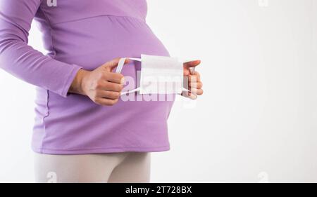 Ein schwangeres Mädchen in einer Fliederbluse hält eine medizinische Schutzmaske vor dem Hintergrund ihres Bauches. Der Begriff des Schutzes gegen Erkältungsinfekten Stockfoto