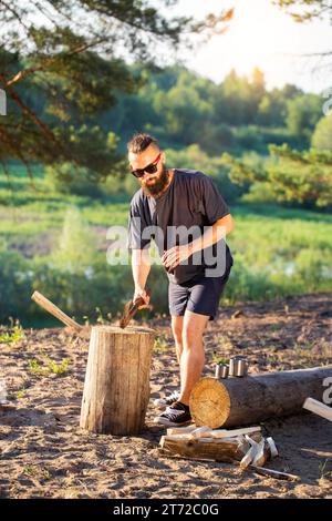 Ein Mann mit einem Bart, ein Holzfäller mit einer Axt in der Hand, schneidet im Sommer Holz in der Natur vor dem Hintergrund eines Waldes und eines Flussufers. Camping Stockfoto