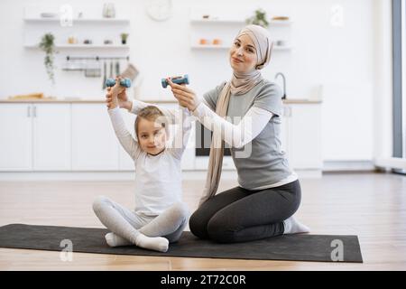 Frau im Hijab hilft süßen Tochter beim Heben von Kurzhanteln auf schwarzer Matte in der Küche. Stockfoto