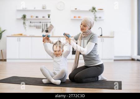 Frau im Hijab hilft süßen Tochter beim Heben von Kurzhanteln auf schwarzer Matte in der Küche. Stockfoto