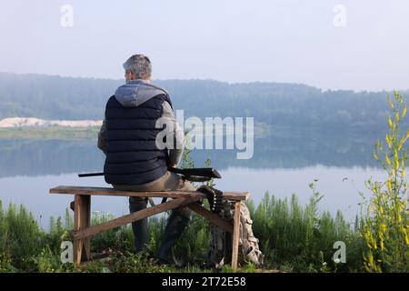 Mann mit Jagdgewehr, sitzt auf einer Holzbank in der Nähe des Sees im Freien, Rückblick. Platz für Text Stockfoto