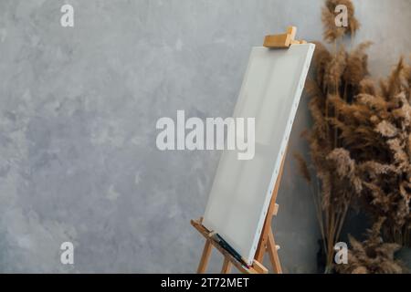 Staffelei mit weißer Leinwand steht auf einem grauen Hintergrund mit braunen Blumen Stockfoto