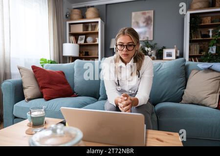 Junge Ärztin, die von zu Hause aus arbeitet, sitzt auf Sofa und hat Videoanruf mit nervösen kranken Patienten, die Menschen hören und sich um sie kümmern. Prof Stockfoto