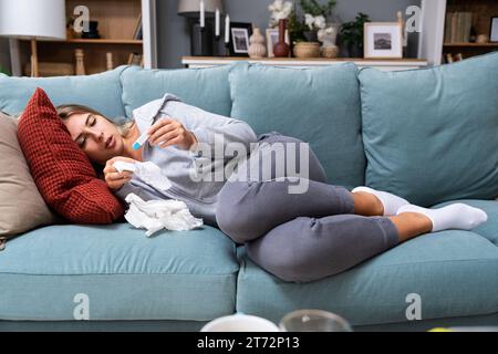 Junge kranke Frau mit Erkältungs- und Grippevirus zu Hause auf dem Sofa, das Körpertemperatur mit einem Thermometer misst, das ihre laufende Nase mit Papiertaschentuch abwischt. Stockfoto