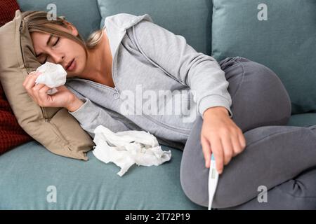 Junge kranke Frau mit Erkältungs- und Grippevirus zu Hause auf dem Sofa, das Körpertemperatur mit einem Thermometer misst, das ihre laufende Nase mit Papiertaschentuch abwischt. Stockfoto