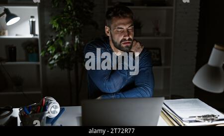 Überfordert und gestresst kämpft der junge hispanische Geschäftsmann mit Rückenschmerzen, der sich in einem schwachen Büro über den Laptop gebeugt hat Stockfoto