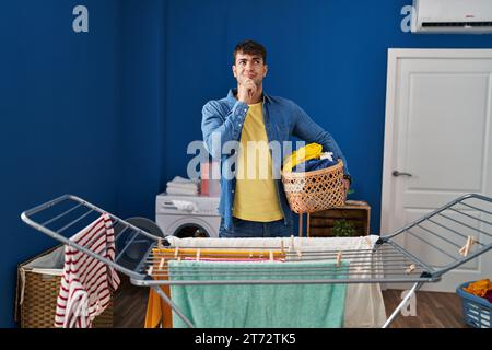 Junger hispanischer Mann, der Kleidung an der Kleidung hängt ernstes Gesicht, denkt mit Hand am Kinn über Fragen nach, nachdenklich über verwirrende Ideen Stockfoto