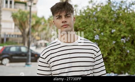 Der junge, gutaussehende hispanische Mann steht, konzentriert sich mit einem ernsten Ausdruck in einem sonnendurchfluteten Park, und sein cooler, entspannter Lebensstil strahlt durch Stockfoto