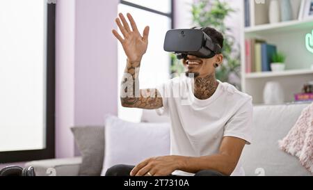 Fröhlich tätowierter junger lateinmann, alle lächeln, chillt auf seinem bequemen Sofa zu Hause, selbstbewusst. Mit virtueller Technik tauchen Sie ein in ein neues Gaming-Level Stockfoto