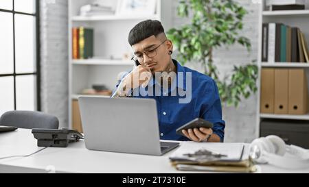 Junger, seriöser lateinmann, der in Gespräche vertieft ist und am Telefon telefoniert, während er im Büro an seinem Laptop arbeitet. Beschäftigter Profi, überarbeiteter empl Stockfoto