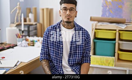 Ernstzunehmender junger lateinmann, tätowierter Künstler in Brille, sitzt drinnen im Kunststudio, vertieft in das Bemalen seiner Leinwand mit Leidenschaft Stockfoto