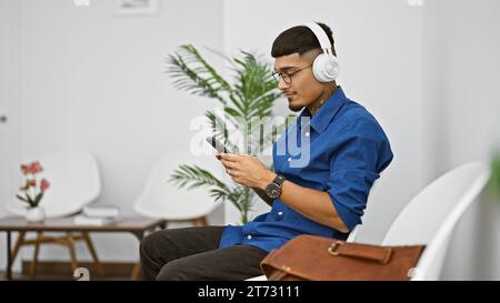 Junger lateinischer Mann, vertieft in sein Telefon, tippte dem Rhythmus, der durch seine Kopfhörer hallt, und auf einem Stuhl im Wartezimmer Stockfoto