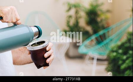 Ein Mann gießt Wasser aus einer Thermoskanne in einen Gefährten. Yerba Mate, traditionelles südamerikanisches Heißgetränk. lateinamerikanischer Mann im mittleren Alter, der draußen genießt. Stockfoto