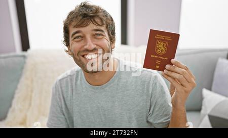 Ein lächelnder junger Mann, der selbstbewusst den finnischen Pass hält, sich zu Hause auf dem Sofa chillt, bereit für sein bevorstehendes Reiseabenteuer! Stockfoto