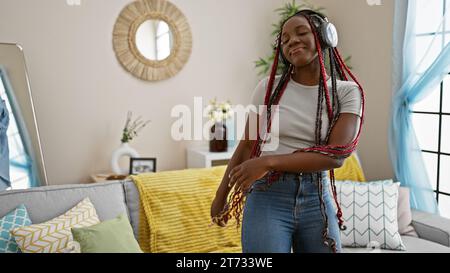 Fröhliche afroamerikanische Frau schaukelte Zöpfe, lauschte Musik, tanzte selbstbewusst im Wohnzimmer, lächelte, während sie ihr entspanntes Leben genießt Stockfoto