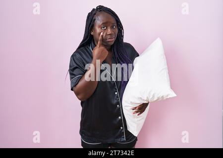 Eine junge afrikanerin, die ein Pijama trägt und ein Kissen trägt, das auf das Auge zeigt und Sie bei einer Geste beobachtet, einem misstrauischen Ausdruck Stockfoto