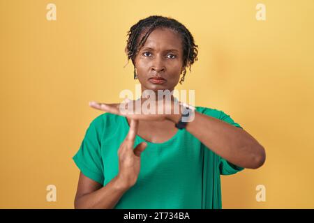 Afrikanische Frau mit Dreadlocks, die über gelbem Hintergrund stehen und mit Händen eine Auszeit-Geste machen, frustriertes und ernstes Gesicht Stockfoto