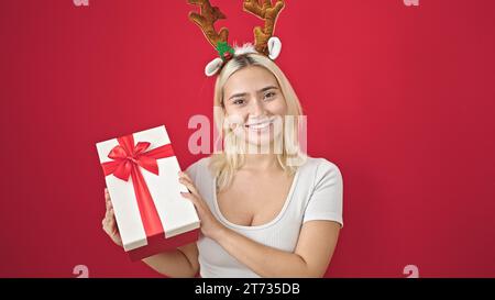 Junge schöne hispanische Frau trägt Rentier weihnachtshut mit Geschenk über isoliertem roten Hintergrund Stockfoto