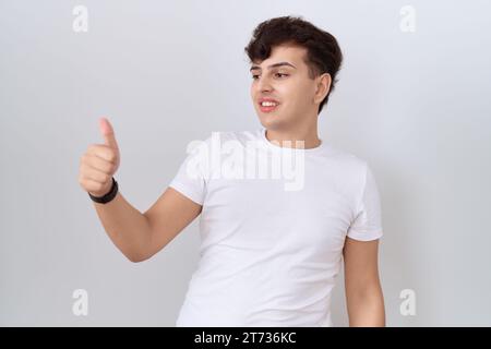 Junger, nicht binärer Mann mit lässigem weißem T-Shirt, der stolz aussieht, lächelnd mit Daumen nach oben Geste zur Seite Stockfoto