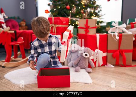 Entzückender hispanischer Junge, der zu Hause am weihnachtsbaum sitzt und ein Geschenk auspackt Stockfoto