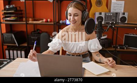 Eine junge Radioreporterin, die im Radio-Studio mit Laptop auf Papier schreibt Stockfoto