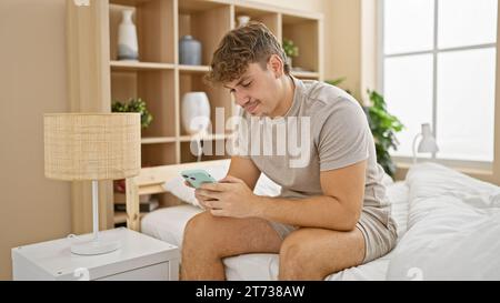 Zu Hause sitzt ein gutaussehender junger hispanischer Mann, völlig in der Zone, morgens bequem entspannt auf seinem Bett, vertieft in sein Handy und tippt ein Chaos Stockfoto