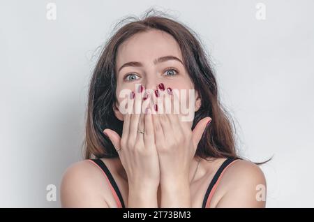 Die überraschte Frau bedeckt ihren Mund mit Händen. Große graue Augen, schwarze Augenbrauen. Helle Maniküre. Stockfoto