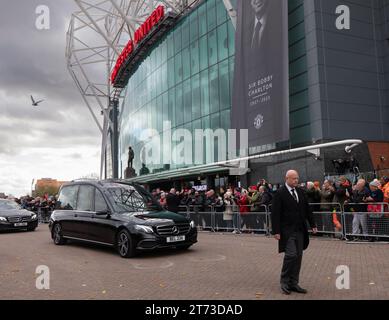 Manchester, Großbritannien. 13. November 2023. Sir Bobby Charlton's Leichenwagen passiert den Old Trafford Football Ground, wo Blumen und Nachrichten an der Gedenkstätte für die Münchner Katastrophe von 1958 hinterlassen wurden. Der Leichenwagen ging an der Trinity Statue vorbei und ging über den Sir Matt Busby Way zum Begräbnis im Stadtzentrum von Manchester. Bild: Garyroberts/worldwidefeatures.com Credit: GaryRobertsphotography/Alamy Live News Stockfoto