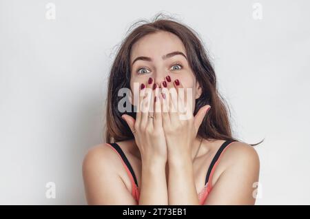 Überraschte dunkelhaarige Frau bedeckt ihren Mund mit Händen. Große graue Augen, schwarze Augenbrauen. Stockfoto