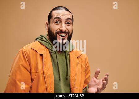 Fröhlicher arabischer Mann mit Bart, der in lässiger Kleidung steht und die Kamera vor beigefarbenem Hintergrund betrachtet Stockfoto