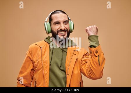 Fröhlicher Mann aus dem Nahen Osten mit Bart, der Musik in kabellosen Kopfhörern vor beigefarbenem Hintergrund hört Stockfoto