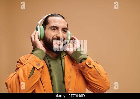 Fröhlicher Mann aus dem Nahen Osten mit Bart, der Musik in kabellosen Kopfhörern vor beigefarbenem Hintergrund hört Stockfoto