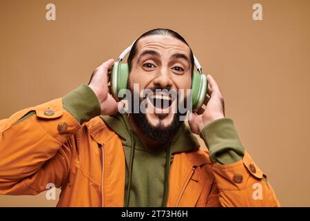 Fröhlicher Mann aus dem Nahen Osten mit Bart, der Musik in kabellosen Kopfhörern vor beigefarbenem Hintergrund hört Stockfoto