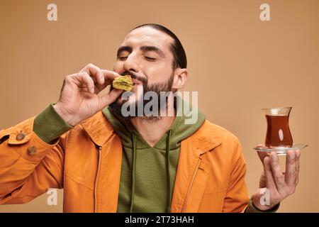 Glücklicher bärtiger Mann, der türkischen Tee in einer traditionellen Glasschale hält und leckeres Baklava auf Beige isst Stockfoto