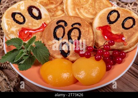 Ein paar Pfannkuchen mit lustigen Gesichtern auf einem Teller mit Pfirsichscheiben, einem Haufen roter Johannisbeeren und Minzblättern Stockfoto