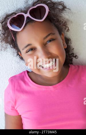 Glückliches lächelndes gemischtes birassisches afroamerikanisches Mädchen Kind mit herzförmiger Sonnenbrille und einem rosa T-Shirt im Urlaub mit einem zahnigen smil Stockfoto