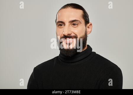 Porträt eines fröhlichen und gutaussehenden arabischen Mannes mit Bart, der in schwarzem Rollkragen vor grauem Hintergrund posiert Stockfoto