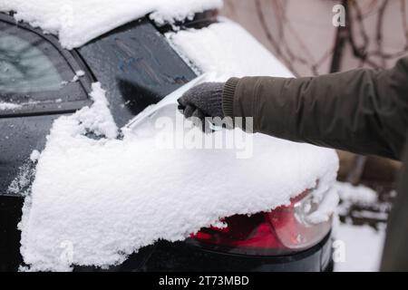 First-Person-Ansicht eines Mannes, der ein schneebedecktes Fenster auf einem Auto mit Schneeschürfer reinigt. Konzentrieren Sie sich auf den Schaber. Kalter Schnee und frostiger Morgen. Schwarzes Auto Stockfoto