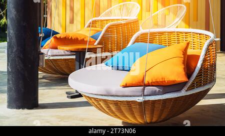 Farbenfrohe Kissen auf einem modernen Tagesbett. Braune Rattan-Liegestühle mit orangefarbenen und blauen Kissen, die mit Seilen auf der entspannenden Terrasse hängen. Sonnenliege zum Sonnenbaden und Stockfoto