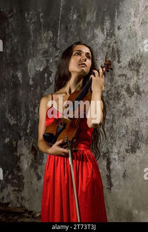 Ein junges Mädchen in einem knallroten Kleid steht an der verbrannten Wand mit einer Geige in den Händen. Stockfoto