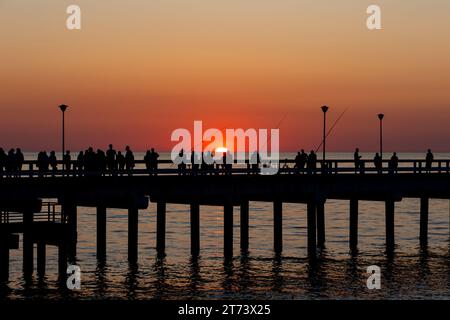 Dunkle Umrisse von Menschen, die während der untergehenden Sonne auf dem Seebrück spazieren gehen. Der Himmel leuchtete orange mit den letzten Sonnenstrahlen. Stockfoto