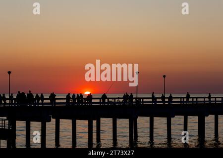 Dunkle Umrisse von Menschen, die während der untergehenden Sonne auf dem Seebrück spazieren gehen. Der Himmel leuchtete orange mit den letzten Sonnenstrahlen. Stockfoto