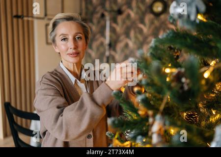 Porträt einer eleganten Frau mittleren Alters hängt Ball an einem Zweig des Weihnachtsbaums in einem gemütlichen Wohnzimmer und blickt in die Kamera. Glückliche weibliche Hausdekoration Stockfoto