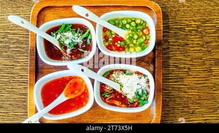 Vier verschiedene scharfe und süße asiatische Gewürze in weißen Schüsseln mit Löffeln ordentlich auf einem rustikalen Holztablett angeordnet. Stockfoto