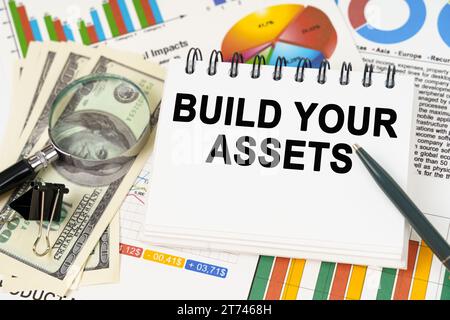 Geschäftskonzept. Auf dem Tisch befinden sich Geschäftsdiagramme, Dollar und ein Notizblock mit der Aufschrift „Build Your Assets“ Stockfoto