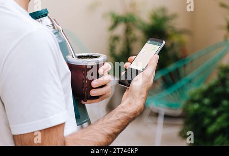 Mann mit Kürbis und Thermos, der ein Handy in der Hand hält, mit einer Chat-App auf dem Bildschirm. Ich schicke Nachricht, während ich Mate trinke. Traditionelles South amer Stockfoto