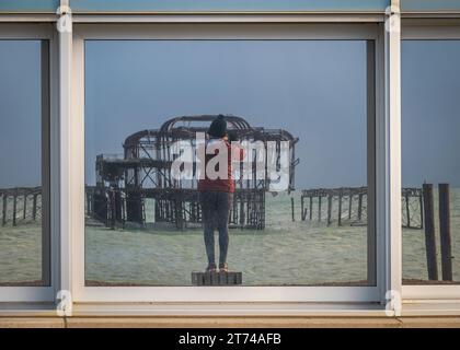 Die Ruinen des West Pier spiegeln sich in den Fenstern des BA i360 an der Küste in Brighton, Sussex, England, wider Stockfoto